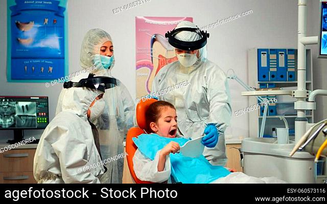 Happy little girl in ppe suit looking in the mirror after dental intervention sitting in new normal stomatological office