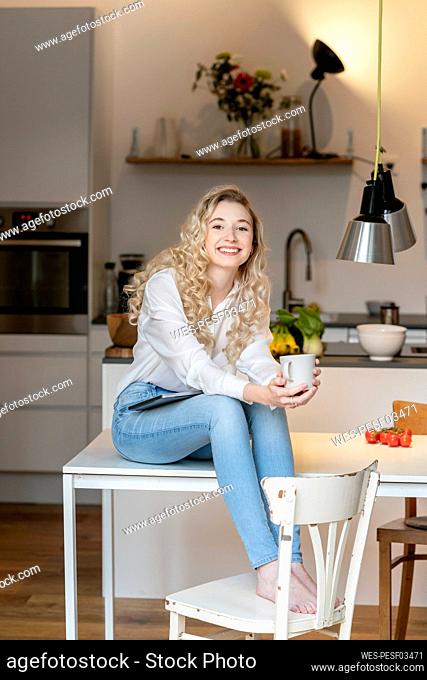 Smiling blond woman holding coffee cup sitting with laptop on table in kitchen at home