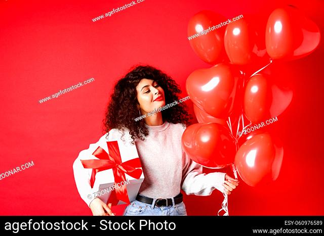 Romantic woman looking dreamy at heart balloons from lover, holding Valentines day gift in cute wrapped box, standing on red background