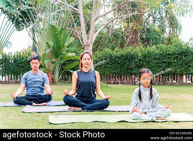 Asian young mother, father practicing doing yoga exercises with child daughter outdoors in meditate pose together in nature a field garden park
