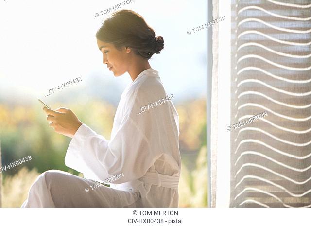 Woman in bathrobe texting with cell phone in sunny doorway