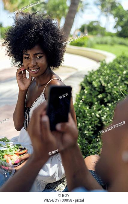 USA, Florida, Miami Beach, young man taking a picture of girlfriend eating a salad in a park