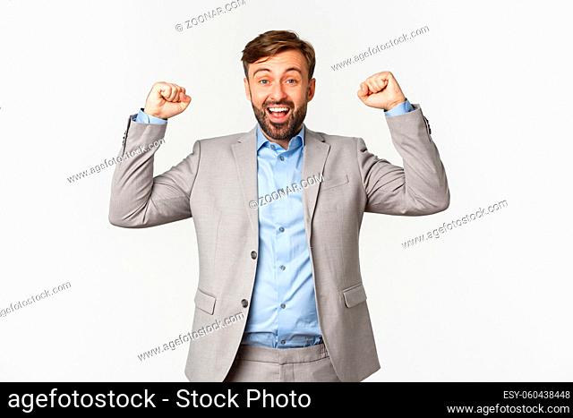 Portrait of relieved and happy bearded man in business suit, rejoicing over win, raising hands up and saying yes with satisfaction