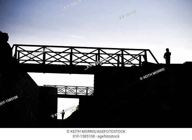 Two seperate people standing, and footbridges, silhouetted at dusk, UK