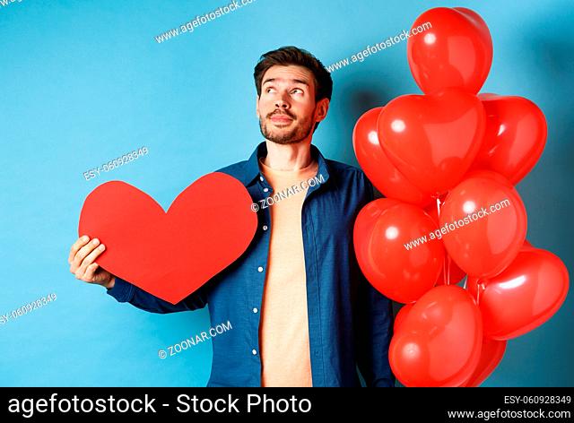 Valentines day and love concept. Man dreaming of soulmate, holding big red heart cutout and balloons, standing over blue background