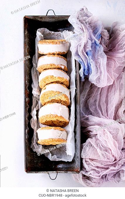 Set of homemade ice cream sandwiches in oat cookies with almond sugar crumbs on baking paper in old metal bowl over gray texture background