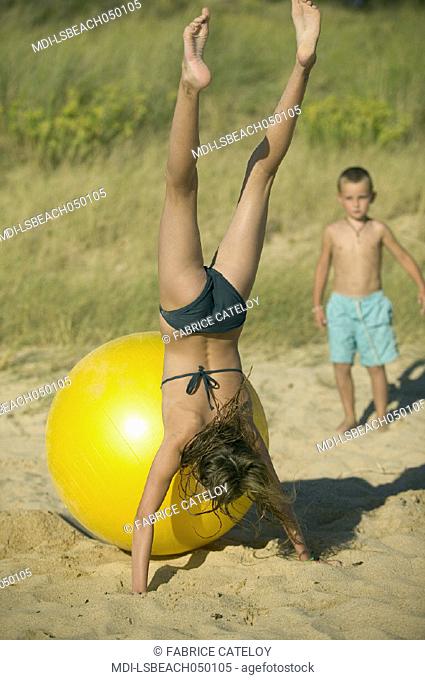 Young girl playing with a big yellow balloon on the sand beach - Little boy looking at her in the background
