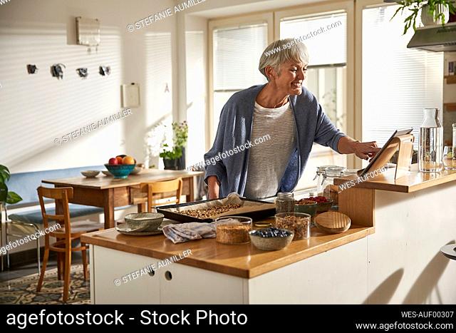 Smiling senior woman using digital tablet in kitchen while preparing granola