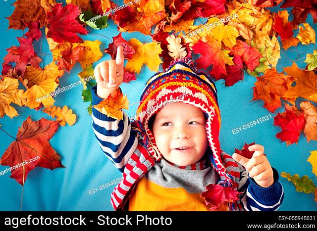 two years old boy dreaming in autumn. Child in hat and scarf playing with maple leaves on blue background
