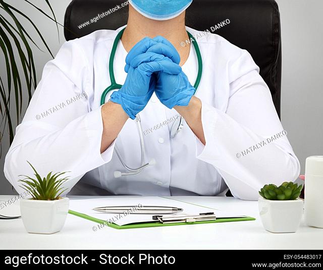 doctor in a white coat and blue latex gloves sits at a white work table in his office, concept of patient reception in the clinic