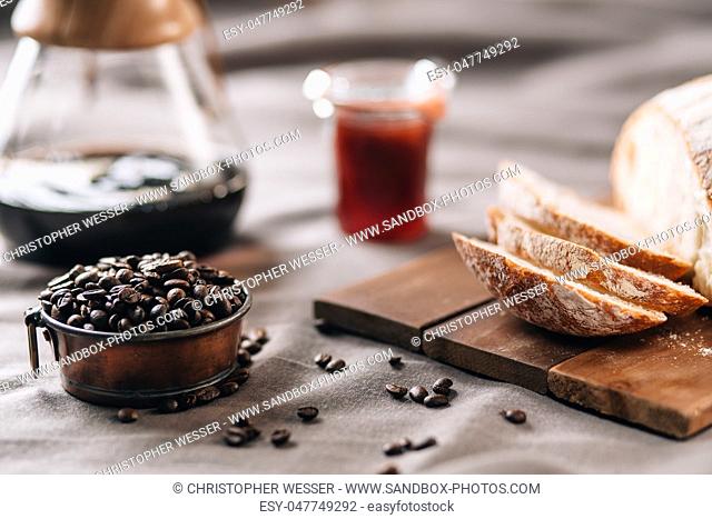 A tabletop scene with coffee beans in a pot, some bread, jam and coffee on a table cloth