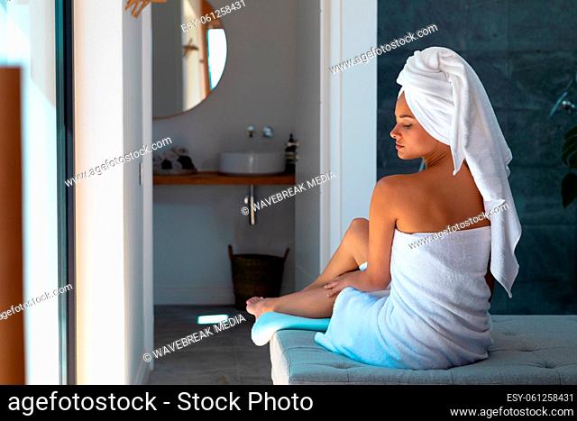 Caucasian woman in a bathrobe applying moisturizer on her leg sitting on the bed at home
