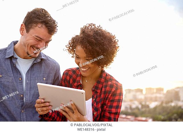 Intimate young couple laughing while looking at the tablet with interest while the sun is setting behind them
