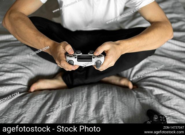 Gamer in white tshirt holding a gamepad in hands and sitting on a couch. Man hands playing a video game with a white controller