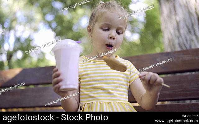 ittle girl eats a colorful gingerbread and holds a milkshake in her hand. Close-up of cute child girl sitting on park bench and eating cookies with a milkshake