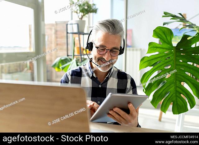 Smiling businessman with headphones and laptop using tablet PC at home