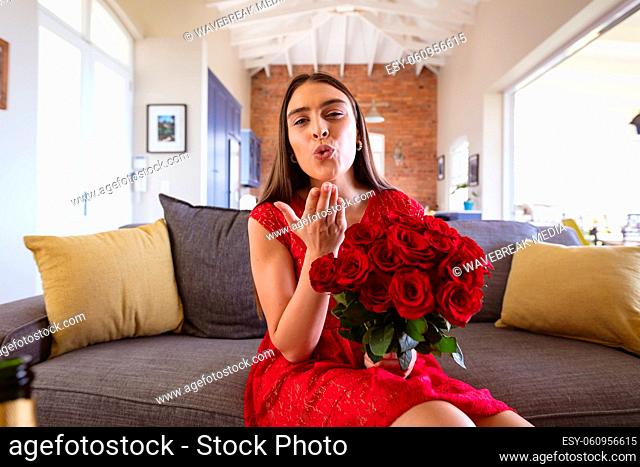 Portrait of biracial woman holding rose bouquet blowing air kiss during virtual date at home