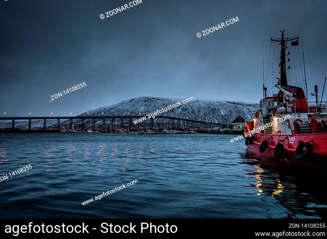 Tromso, Norway - December 2018 : Fishing boat in port and harbour in Tromso, with famous Tromso Bridge across Tromsoysundet strait in the background