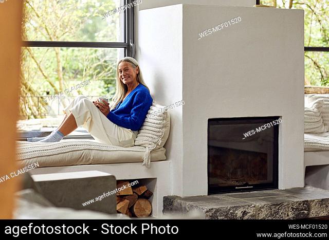 Smiling mature woman with coffee cup sitting on window seat at home