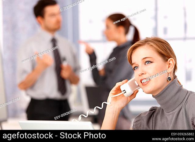 Young businesswoman talking on phone in office, colleagues chatting in background