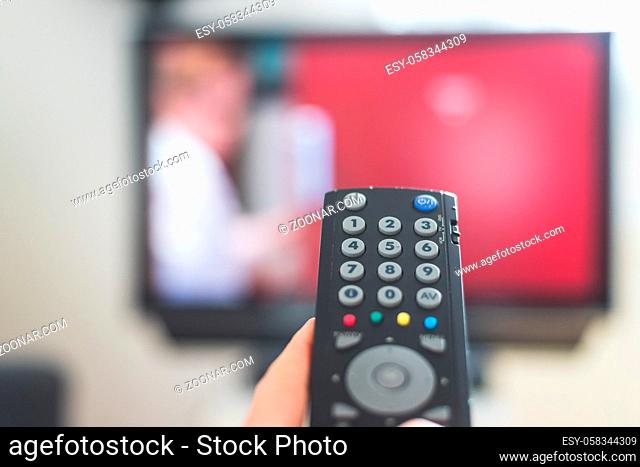 Holding a TV remote control in the hand, foreground, tv in the blurry background. Streaming