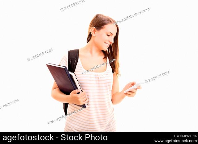 Beautiful young student girl with a backpack, holding notebooks, smiling and looking at her phone, isolated on white background