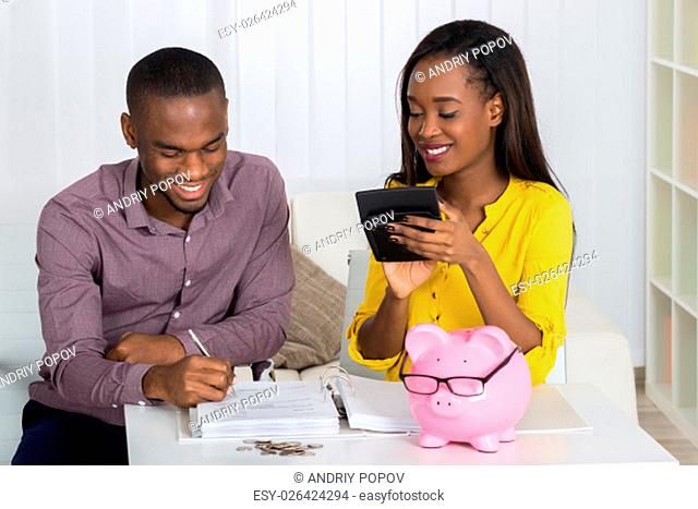 Happy Young Couple Calculating Bill With Coins And Piggybank On Desk