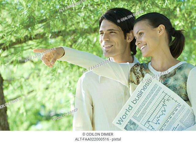Couple standing outdoors holding newspaper, woman pointing, both looking out of frame