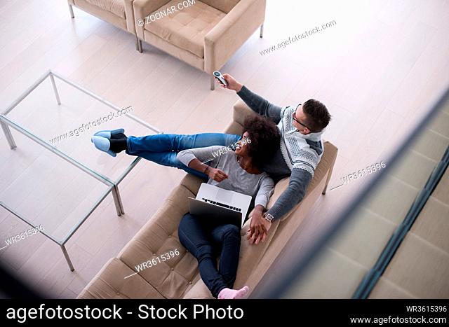 Young multiethnic couple relaxing on the sofa in the luxury living room, using a laptop and remote control