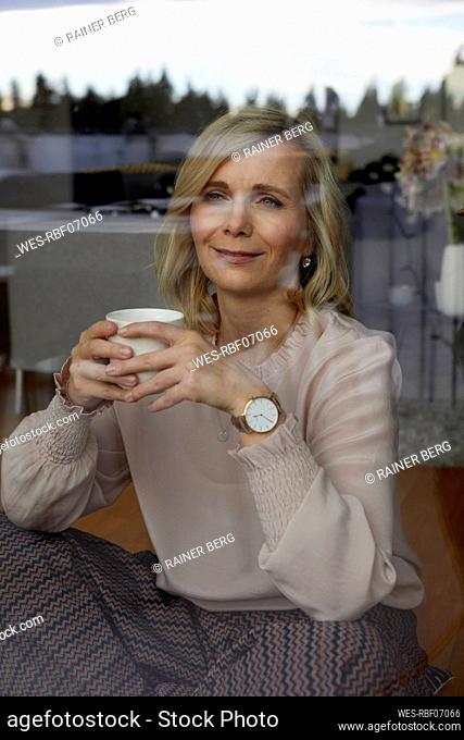 Smiling blond woman holding coffee cup behind windowpane at home