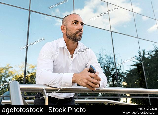 Man holding smart phone leaning on railing in front of building