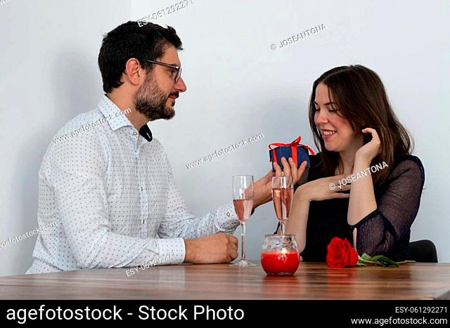 young guy giving a gift with a red ribbon to his girlfriend on valentine's day, with champagne candle and roses