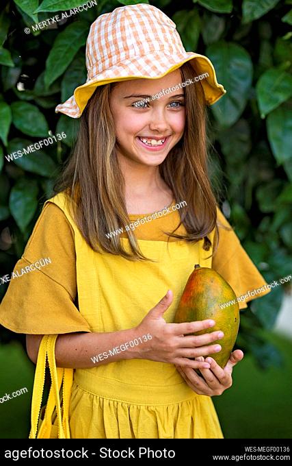 Happy girl wearing hat standing with mango fruit in front of plant