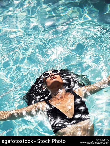 Serene, sensual woman in sunglasses and bikini floating in sunny swimming pool