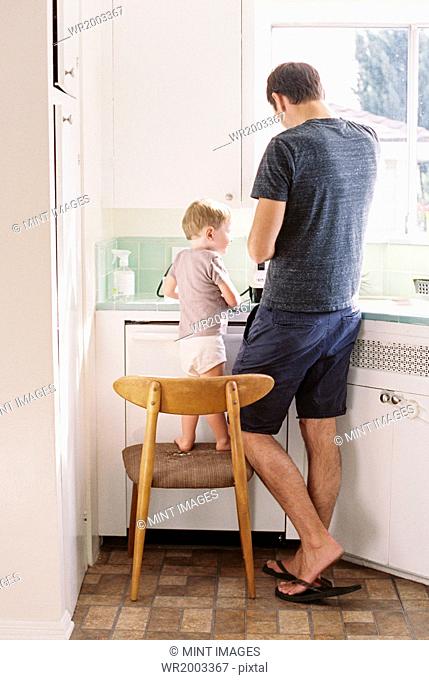 Man standing in a kitchen, his son standing on a chair beside him