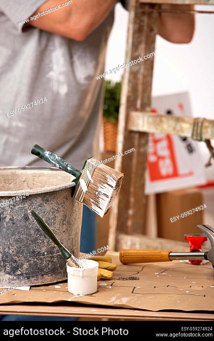 Focus on painting equipment, brush and bucket, man standing by ladder in background