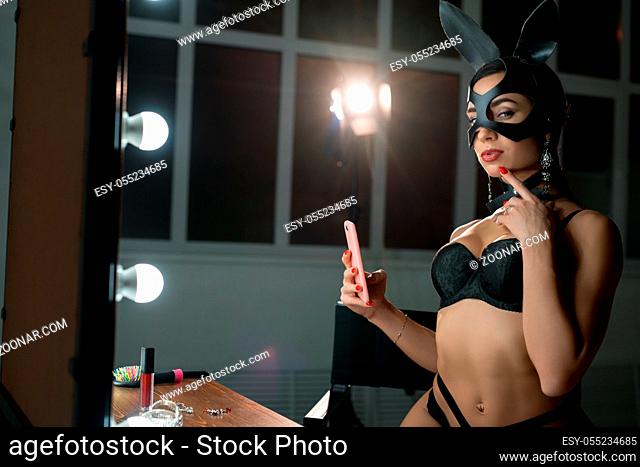 Girl in mask and bunny ears wearing black lingerie sitting on a chair and making selfie in the mirror view Girl in mask and bunny ears wearing black lingerie...