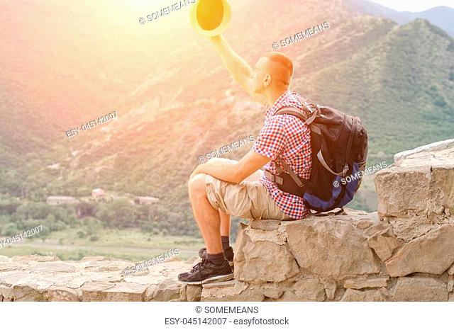 Young man with backpack sits on stone wall with raised hat against the background of green mountains