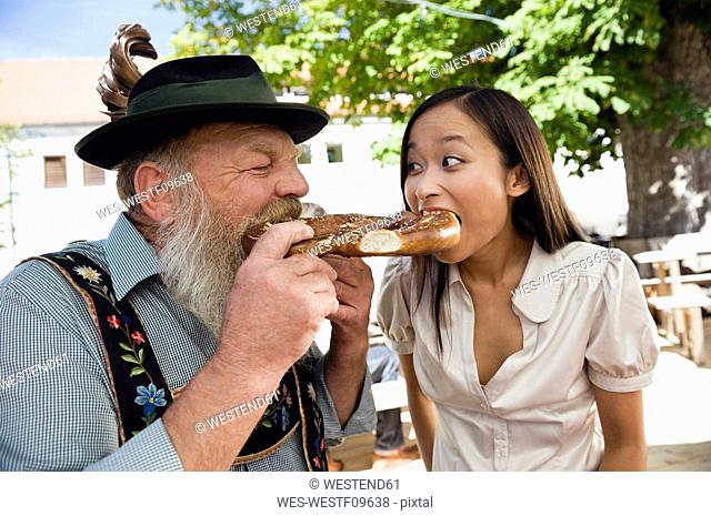 Germany, Bavaria, Upper Bavaria, Bavarian man and Asian woman in beer garden eating pretzel, portrait