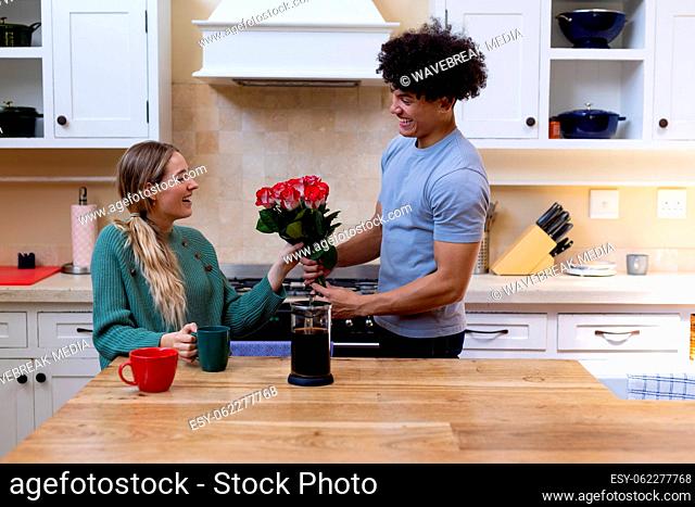 Biracial young husband giving red roses to happy wife having coffee at table in kitchen