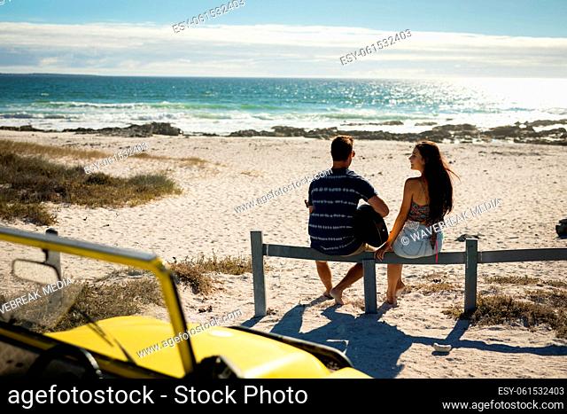 Happy caucasian couple sitting on beach by the sea playing guitar