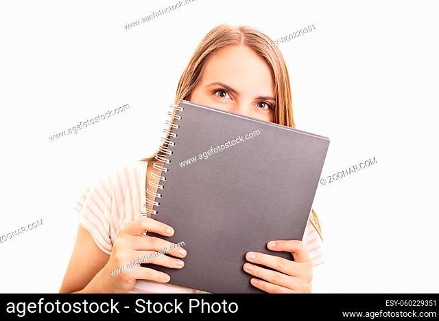 Beautiful girl hiding her smile behind a notebook, isolated on white background. Girl with a mischievous look hiding behind her notebook