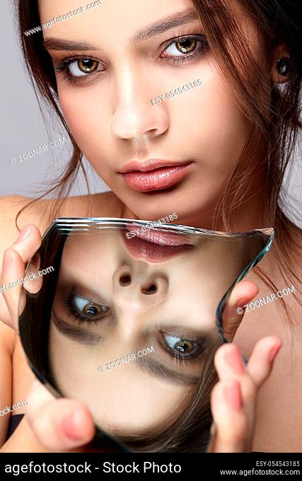 Girl with a shard of the mirror. Female with mirror shard in hand posing on gray background. Face reflection in mirror splinter