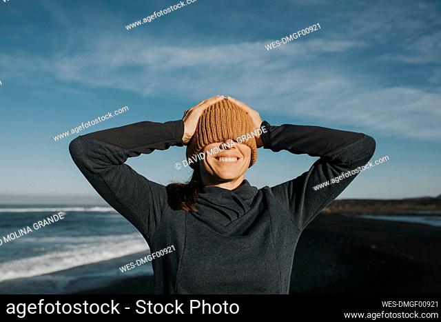 Happy woman covering eyes with knit hat in front of sky