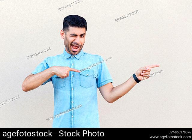 Portrait of funny handsome young bearded man in blue shirt standing, winking, making crazy face, looking at camera, pointing at left side