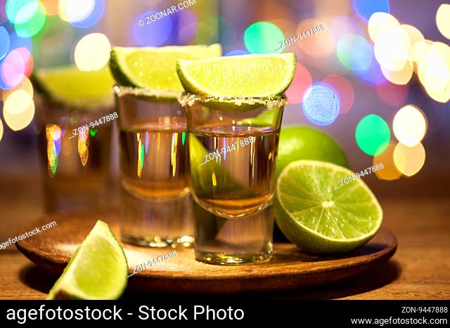 Gold tequila shots with lime fruits on wooden table on bar lights background