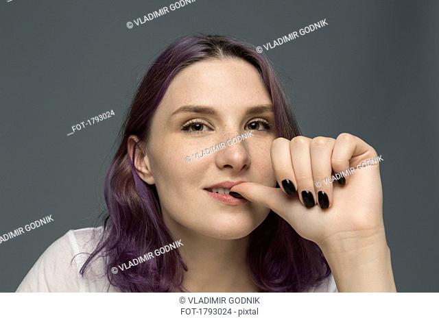 Portrait of a young woman with dyed hair biting nail against gray background