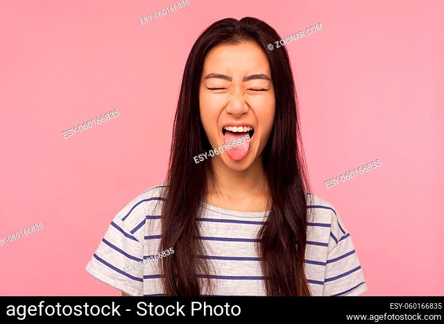 Portrait of funny girl with long hair in striped t-shirt showing tongue and keeping eyes closed with naughty childish grimace, expressing disobedience