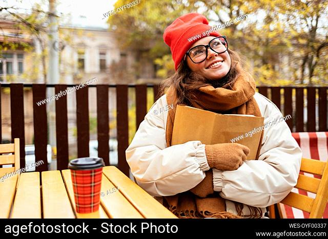 Thoughtful woman smiling while embracing book sitting at sidewalk cafe