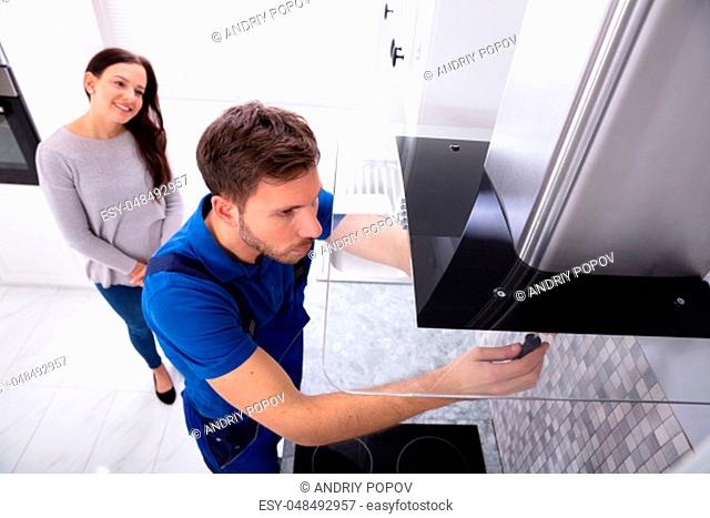 Smiling Woman Standing In Front Of Male Worker Fixing Kitchen Hood With Screwdriver